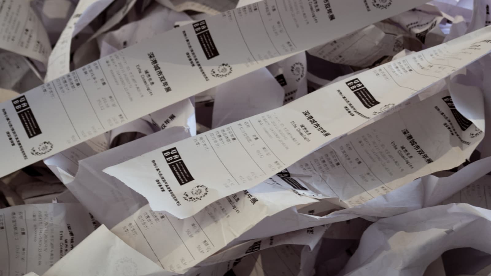 Editorial photograph of a tall stack of identical white paper receipts beside a single elegant hand-bound leather journal on a dark slate surface.